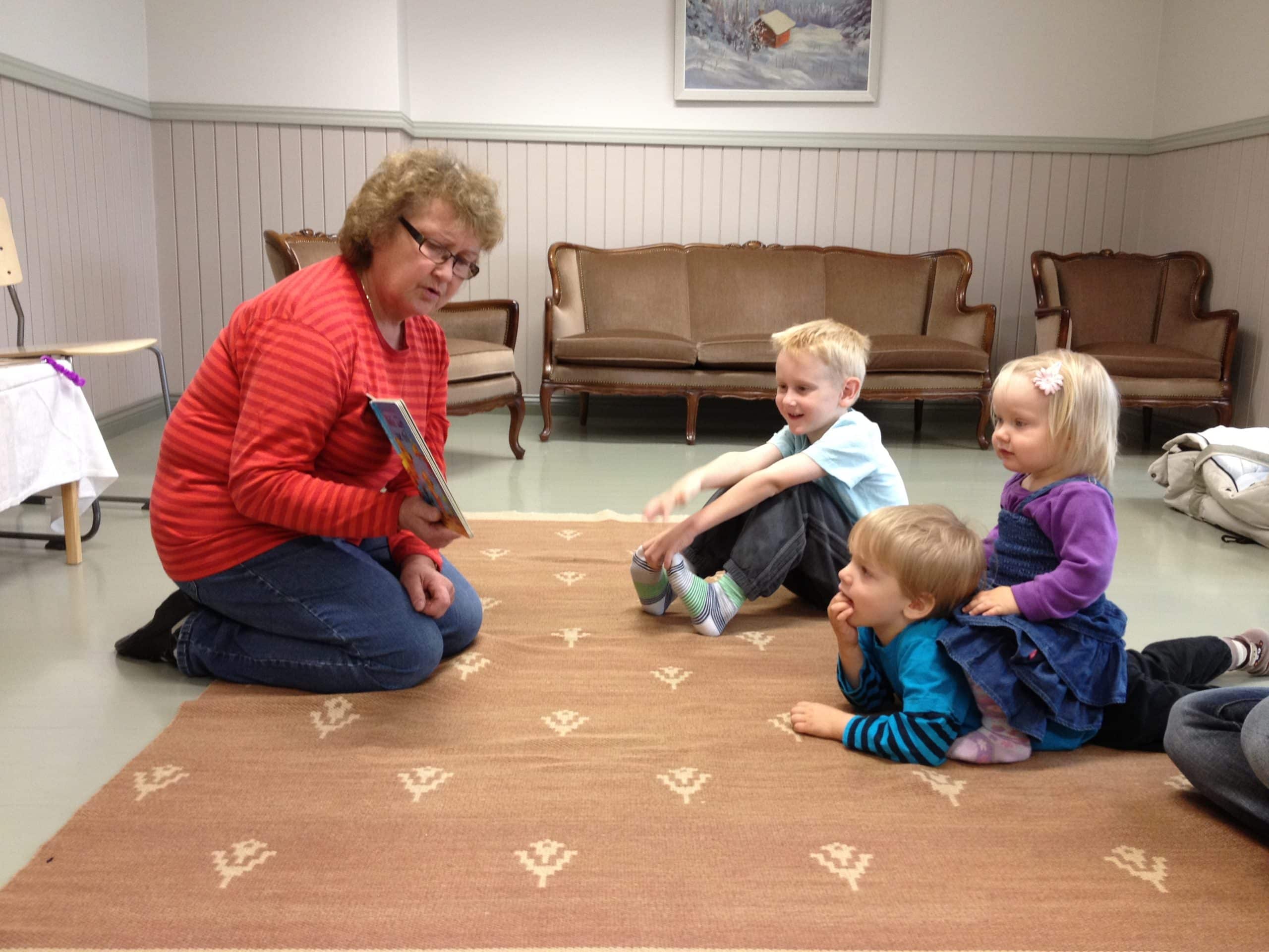 Ohjaaja Aila Vaskela lukee lapsille. Venla kuuntelee satua turvallisesti isoveljensä Akselin kyydissä. Taustalla Aleksi.